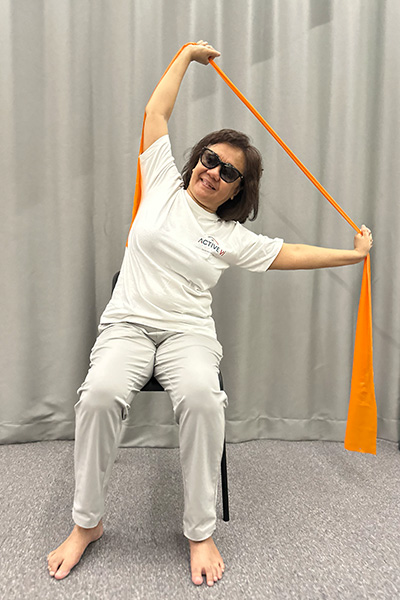 A smiling visually impaired woman seated on a chair performs a side stretch using an orange resistance band. She holds one end of the band with her right arm raised straight up, and the other end with her left arm extended out to the side.