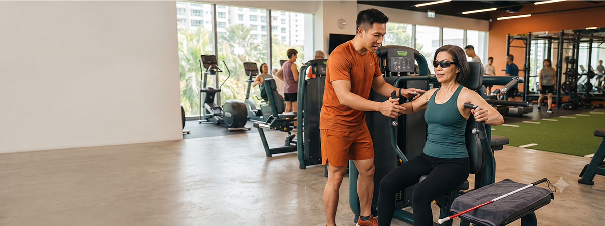 A female gym-goer wearing dark glasses receives verbal guidance from an instructor while using a seated chest press machine. A folded white cane is on a towel nearby.
