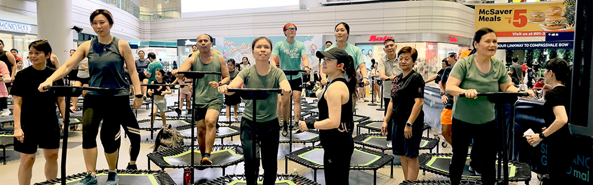 A diverse group of adults participating in a fitness class on individual hexagonal mini-trampolines with handlebars, located in a brightly lit indoor shopping mall atrium.