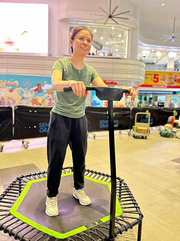 Pei Pei smiling while standing on a mini fitness trampoline, holding onto the front handlebar for support.