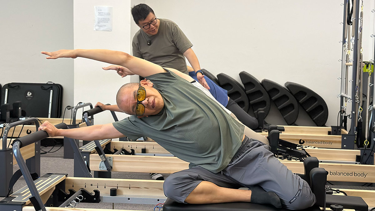 A visually impaired man wearing yellow-tinted glasses performs a side stretch on a Pilates reformer machine while an instructor observes closely from behind.