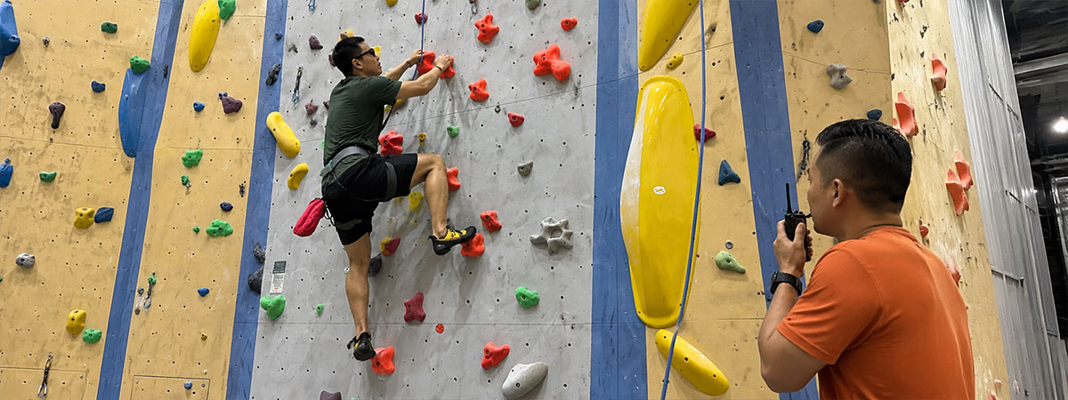 A visually impaired climber scales an indoor climbing wall while a guide below provides spoken directions and manages the safety rope.