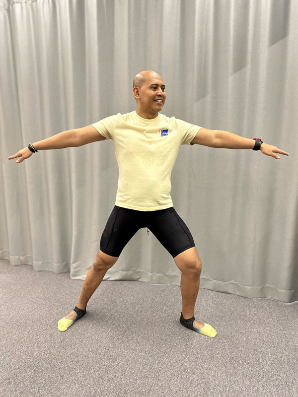 A smiling man performs the yoga Warrior II pose indoors. He stands with his legs wide apart, his right knee bent, and both arms extended straight out to his sides parallel to the floor, looking outward over his right hand.