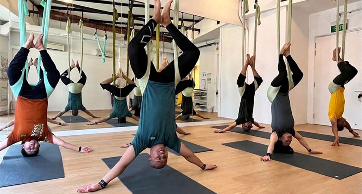 A diverse group of participants hang upside down in hammocks during an inclusive aerial yoga stretch session inside a bright, mirrored studio.