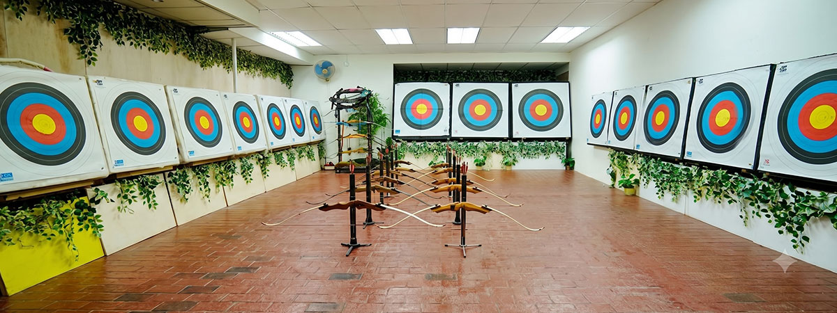 An empty, well-lit indoor archery range featuring multiple lanes of large-format multi-colored targets and organized recurve bows on stands, ready for a recreational session.