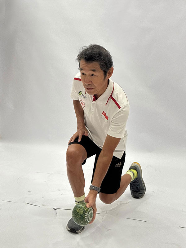 A man in a white Singapore Disability Sports Council polo shirt and black shorts kneels on one knee against a white studio backdrop. He looks focused straight ahead while holding a green, speckled lawn bowl close to the ground, posed in a stance ready to roll it.