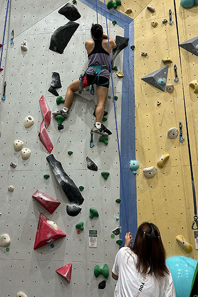 A visually impaired climber scales a tall indoor rock wall, attached to a safety rope. Below, a sighted guide stands looking up, holding the rope and providing verbal instructions.