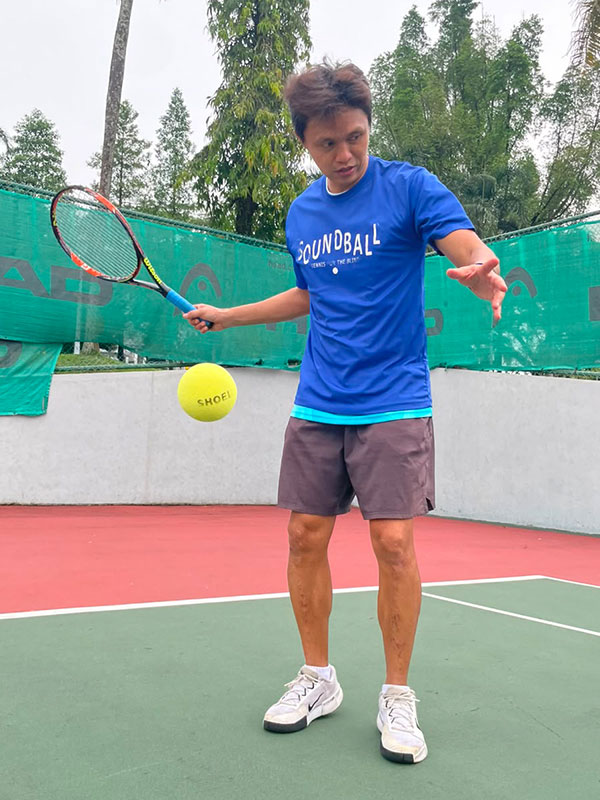A man wearing a blue t-shirt that says "SOUNDBALL: Tennis for the Blind" stands on an outdoor tennis court. He has his right arm extended holding a tennis racquet and his left hand open, having just dropped a large, bright yellow sponge tennis ball. He is looking intently at the ball as it falls mid-air. A green fence and tall palm trees are visible in the background.