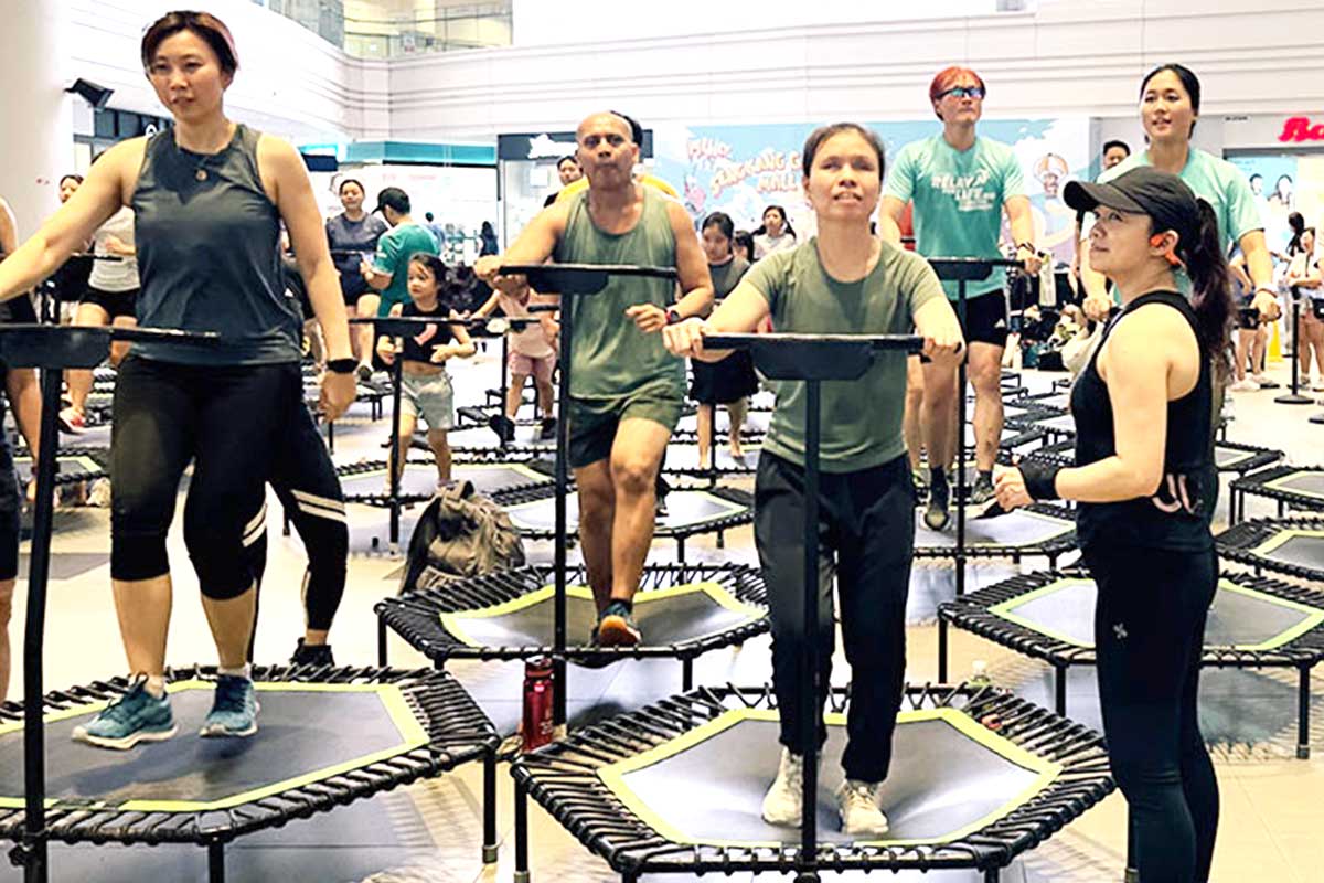 A visually impaired woman smiles while exercising on a mini-trampoline holding a stabilising bar, guided closely by a female volunteer standing right next to her. They are surrounded by other smiling participants enjoying an inclusive group jumping fitness session in a bright, spacious indoor atrium.