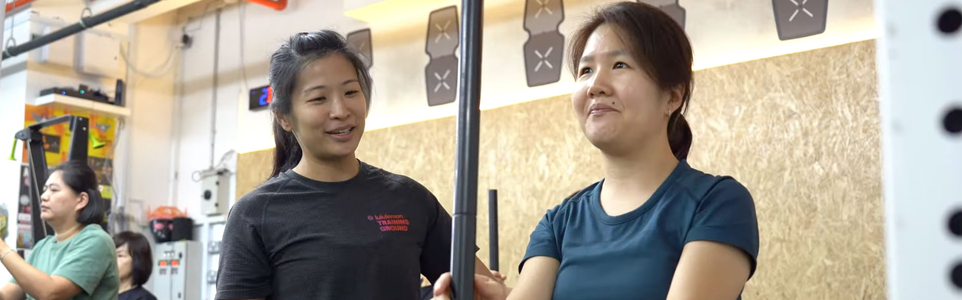 A fitness volunteer smiling and guiding a person with vision impairment as they hold a workout bar during an indoor exercise session.