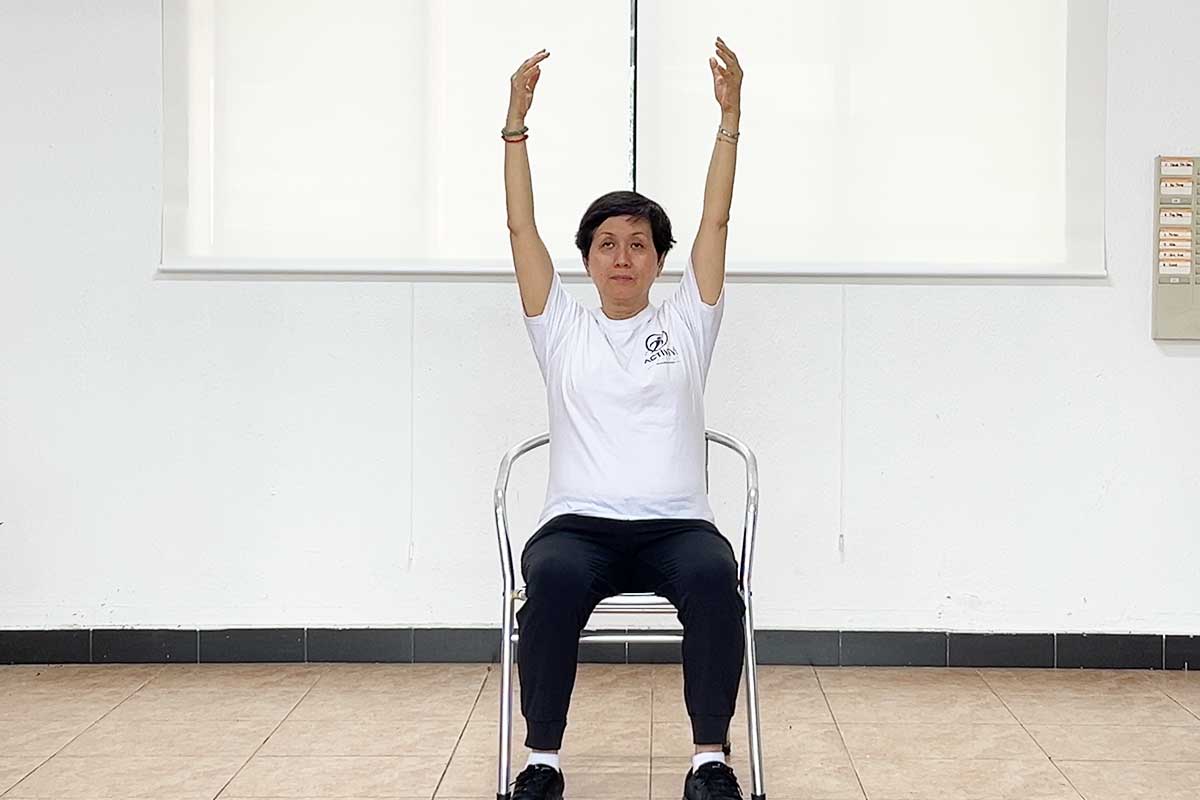 An Asian woman with short black hair sits on a white chair, facing forward in a bright, white room. She wears a white t-shirt featuring the ACTIVE VI logo on the left chest, paired with black track pants and black sneakers. Her arms are raised straight up toward the ceiling. The floor consists of light brown, square tiles, and behind her is a plain white wall with horizontal paneling. A wall-mounted electrical panel is visible on the right side of the background.